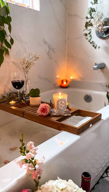 reviewer photo of bath caddy set up, with lit candle, glass of wine, heart-shaped succulent, and flowers