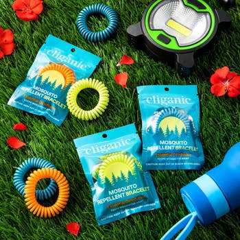 Cliganic Mosquito Repellent Bracelets, three colorful bracelets and their packaging shown on grass with other camping gear, including a LED light and water bottle