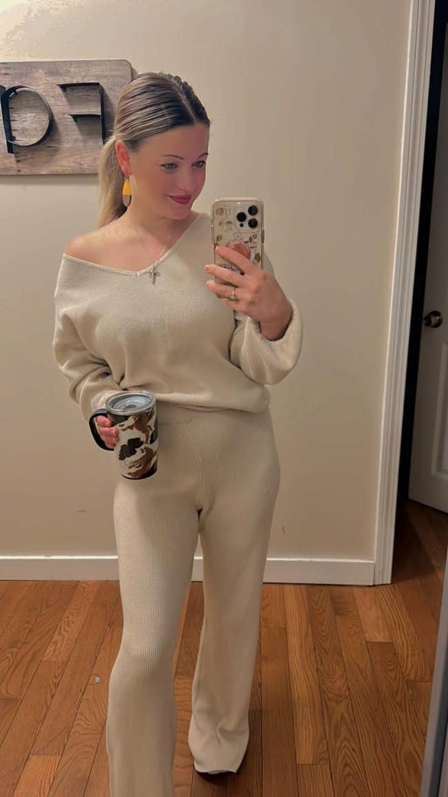 Person taking a mirror selfie wearing a cozy knit lounge set, holding a patterned mug, with a modern wooden wall decoration in the background