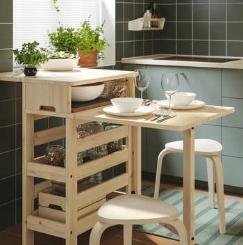 Compact room  setup with a woody  array  and 2  stools, acceptable   for 2  eating  with dishware and vino  glasses, surrounded by greenish  tiled walls and plants