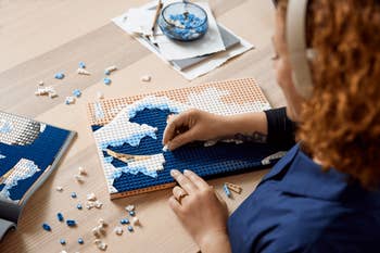 Person assembling mosaic art with small tiles on a table, focused on intricate blue and white sections near an instruction booklet