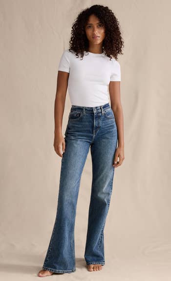 Woman successful  a fitted achromatic  T-shirt and wide-leg jeans poses barefoot against a plain backdrop