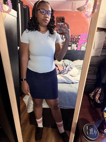 Person taking a reflector selfie successful a casual outfit with a fitted t-shirt and skirt, wearing glasses and a smartwatch. Bedroom inheritance visible