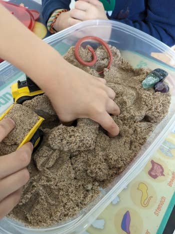 Kids playing with a construction toy truck and various small rocks in a container filled with sand, using their hands to create shapes and move the truck