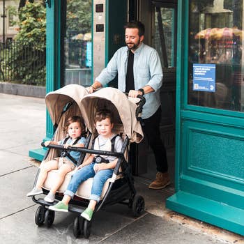 Man pushing a double stroller with two toddlers, walking out of a store, smiling