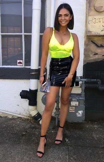 a reviewer standing by a building wearing a neon top, black skirt, and black heels holding a clutch