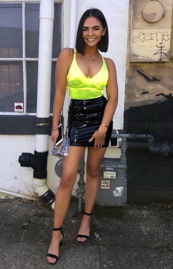 a reviewer standing by a building wearing a neon top, black skirt, and black heels holding a clutch