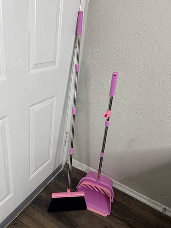 An pink broom with a pink dustpan with slotted edges to clean the bristles of the broom 