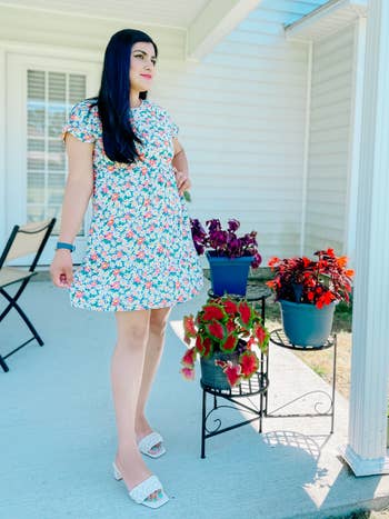 reviewer in a floral dress and white sandals standing on a porch with potted flowers nearby