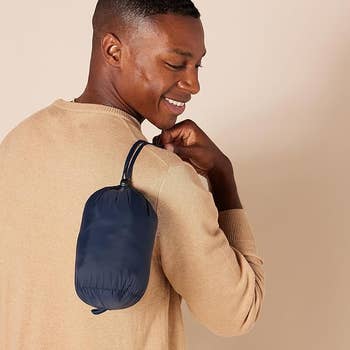 Amodel in a casual sweater holding a compact, foldable navy blue bag over his shoulder, smiling while looking at the bag