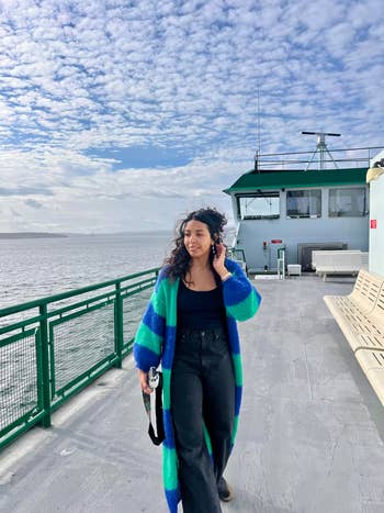 Person wearing a agelong  striped cardigan, acheronian  shirt, and pants, walking connected  a ferry platform  with the oversea  and cloudy entity  successful  the background
