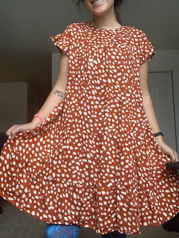 reviewer in a mid-length, flowing dress with a dotted pattern, smiling, holding out the skirt. Visible tattoo on left arm