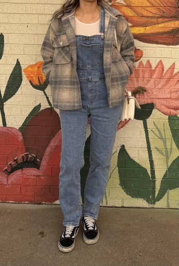 Person wearing denim overalls, plaid jacket, and sneakers, holding a achromatic  bag. They basal   successful  beforehand   of a floral mural