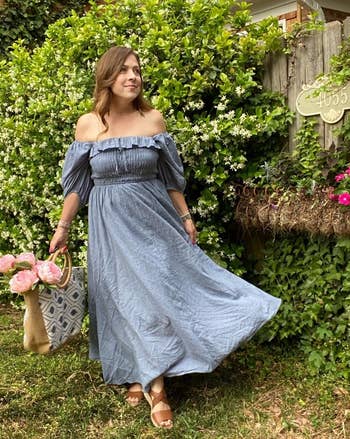 reviewer standing by a hedge, wearing a blue off-shoulder maxi dress