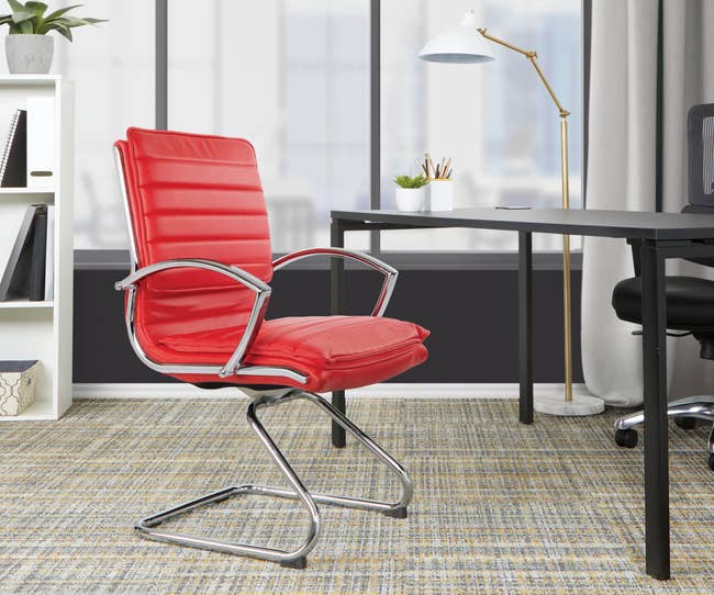 Red leather chair with silver armrests and sled base on a carpet in front of black desk and windows