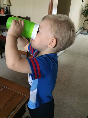 Child drinking from a large green cup indoors, standing by a table