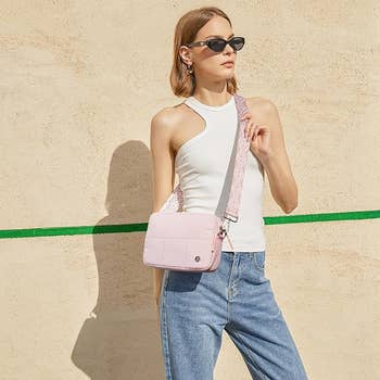 Person in a sleeveless white top and jeans poses with a crossbody bag, wearing sunglasses, against a plain background
