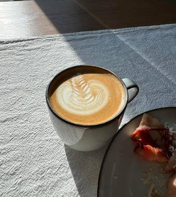 Latte with art on a table next to a partial view of a breakfast plate, in a sunny setting. Ideal for morning shopping inspiration