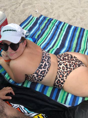 Person lounging on a striped beach towel, wearing a leopard print swimsuit, sunglasses, and a white cap