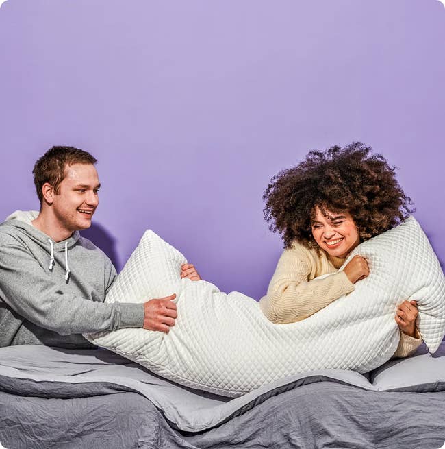 Two radical   connected  a furniture  holding a large, textured pillow, smiling. They are wearing casual, comfy  sweaters. The inheritance  is plain
