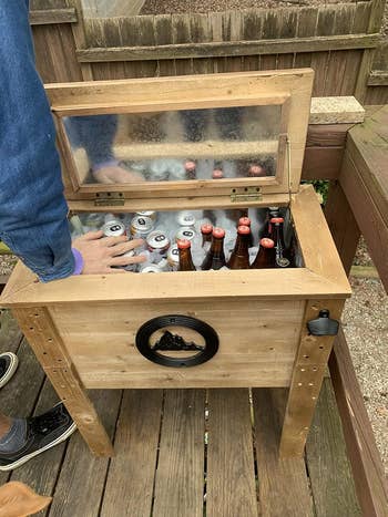 Person reaching into a rustic wooden cooler filled with various cans and bottles on a wooden deck