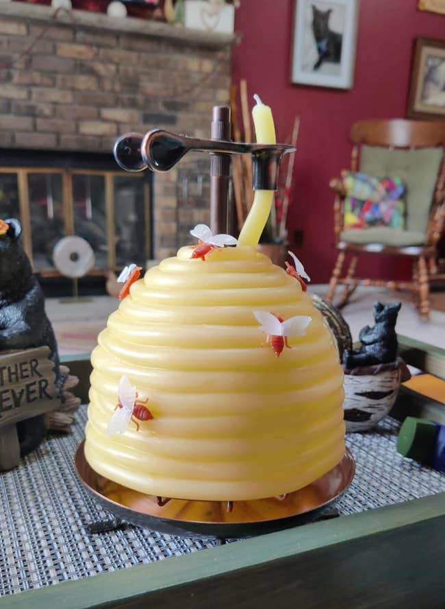 A beehive-shaped candle with small decorative bees and a bent wick is displayed on a table in a living room with a rustic brick fireplace in the background