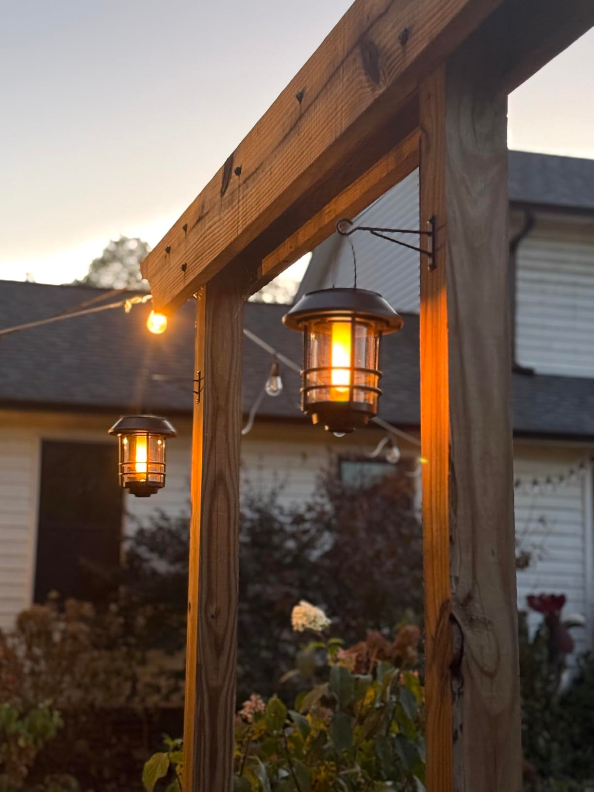 Hanging outdoor lanterns connected  a woody  pergola beam, illuminated astatine  dusk, with a location   and plot  successful  the background. Ideal for patio decor