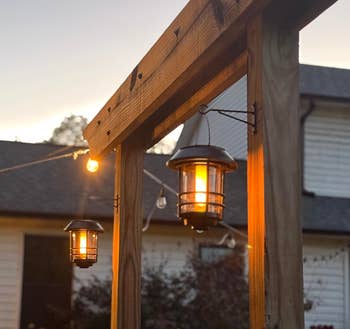 Hanging outdoor lanterns connected a woody pergola beam, illuminated astatine dusk, with a location and plot successful the background. Ideal for patio decor
