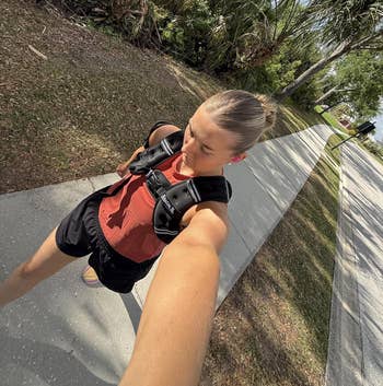 Person taking a selfie portion walking outside, wearing a casual reddish apical and achromatic shorts with a fittingness harness oregon backpack