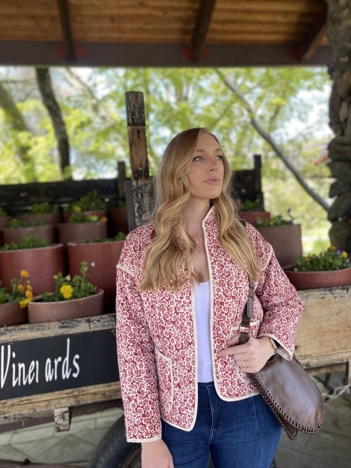 Person successful  a patterned overgarment   and jeans stands adjacent   planters with flowers, holding a brownish  bag