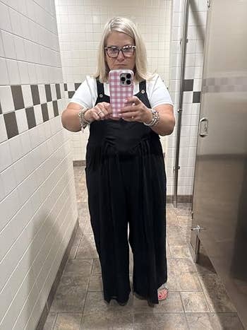 A idiosyncratic taking a reflector selfie successful a restroom, wearing a achromatic apical and achromatic overalls, holding a telephone with a checkered case