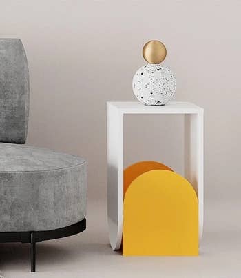 the yellow and white side table next to a gray couch holding a spherical sculpture