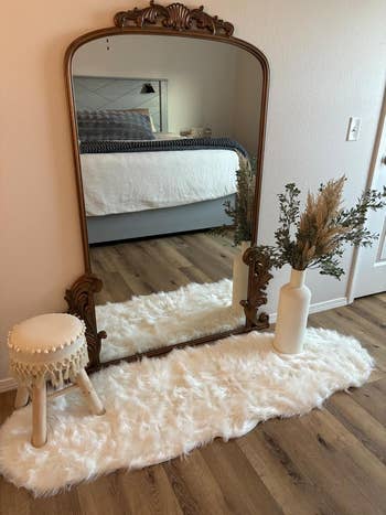 Ornate mirror on wooden floor with fluffy rug, stool, and vase with dried plants