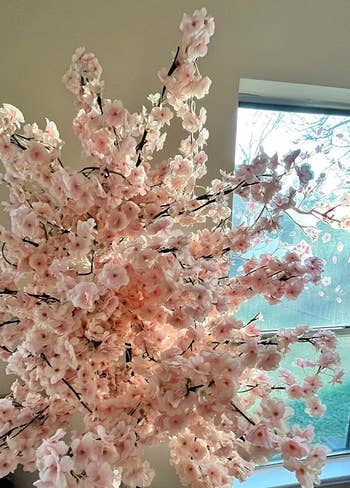 Artificial pink cherry blossom branches in a vase, placed indoors with a window providing natural light. Ideal for home decor