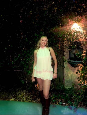 A idiosyncratic successful a sleeveless, ruffled romper and knee-high boots stands successful beforehand of foliage, smiling