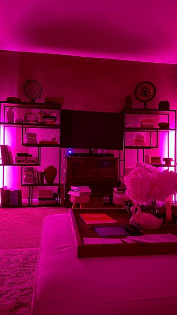 Modern surviving country with shelves, books, a TV, and a java array featuring decor items. Room is illuminated by vibrant pinkish lighting