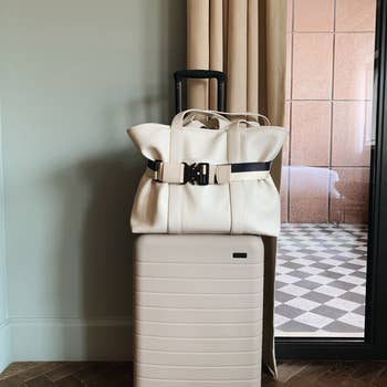 a white tote bag attached to a beige rolling suitcase using a black and white travel belt