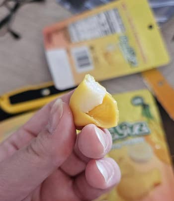 A close-up of a hand holding a bitten-into candy, showing its chewy outer layer and creamy filling. Packaging in the background hints the candy is Asian in origin