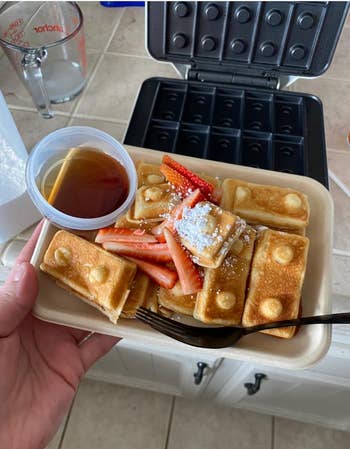 Rectangular waffles with powdered sugar and strawberries, served with syrup in a cup. Waffle maker in the background