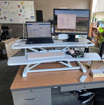 A white two tiered desk riser holding a keyboard on one shelf and a laptop and monitor on the higher one above it 