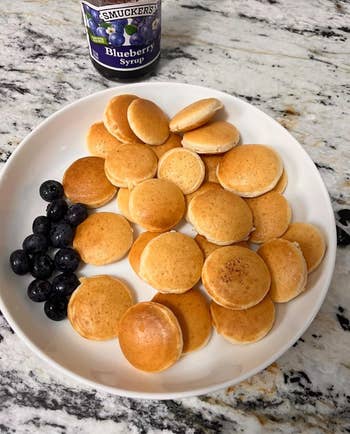 Small pancakes with blueberries on a plate, accompanied by a jar of Smucker's blueberry syrup, set on a marble countertop