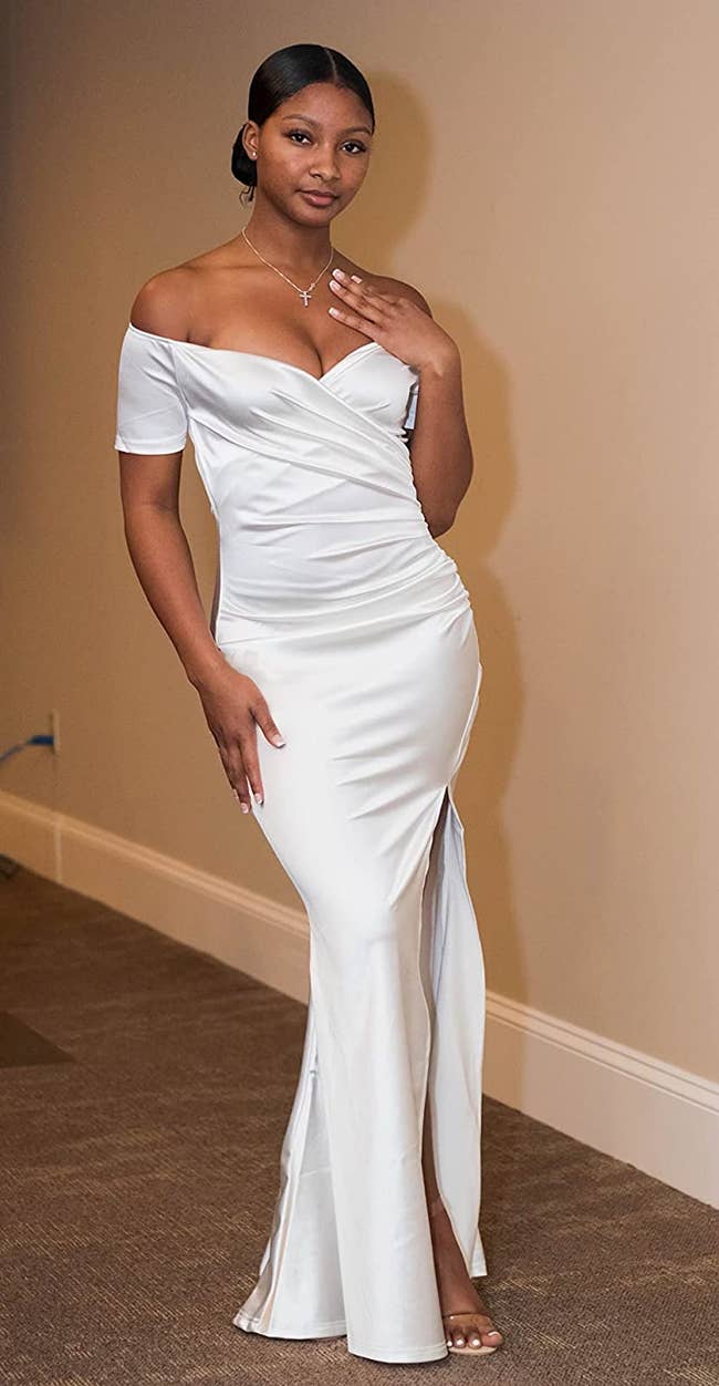Reviewer wearing floor length white formal off the shoulder dress with front leg slit and criss-cross design in front of white wall