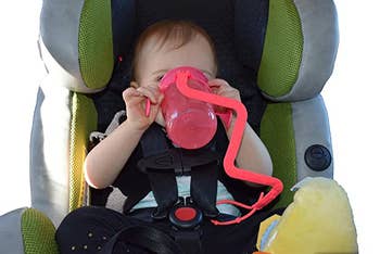 A baby drinking from a sippy cup that is attached to their car seat buckle by the strap