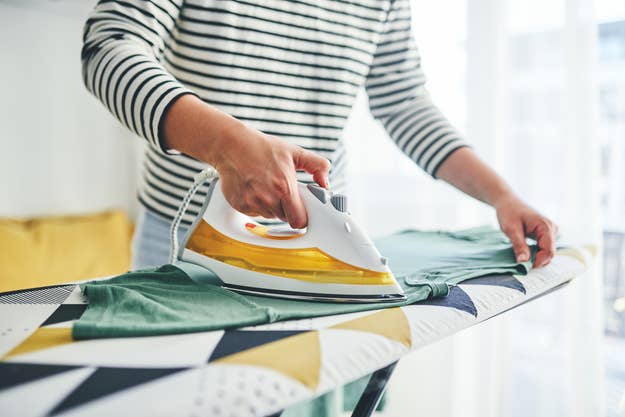 A person wearing a striped shirt is ironing a green garment on a patterned ironing board at home