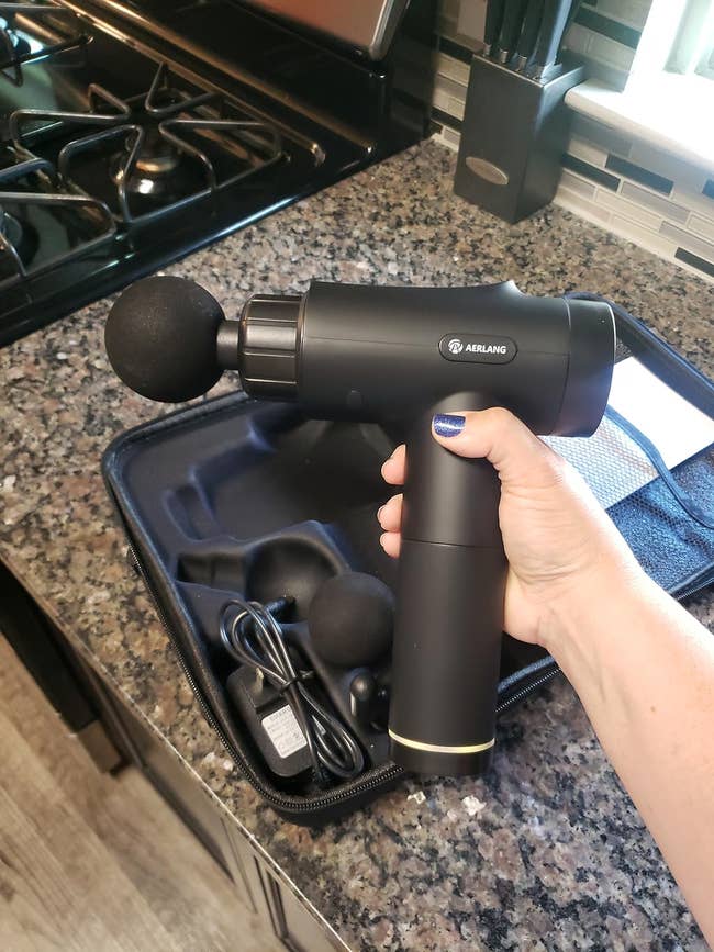 Hand holding a percussion massage gun with accessories on a kitchen countertop, suitable for muscle relaxation and recovery