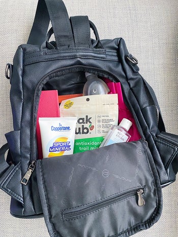 BuzzFeeder's photo of the black faux leather backpack holding snacks, sunscreen, an umbrella, glasses case, water bottle, and hand sanitizer in its theft-proof back pocket