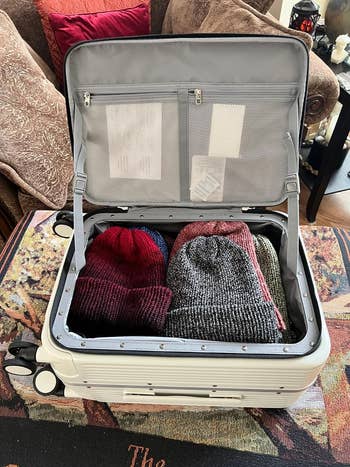 a reviewer's open suitcase with neatly packed clothes, including folded knit hats, on a patterned rug indoors