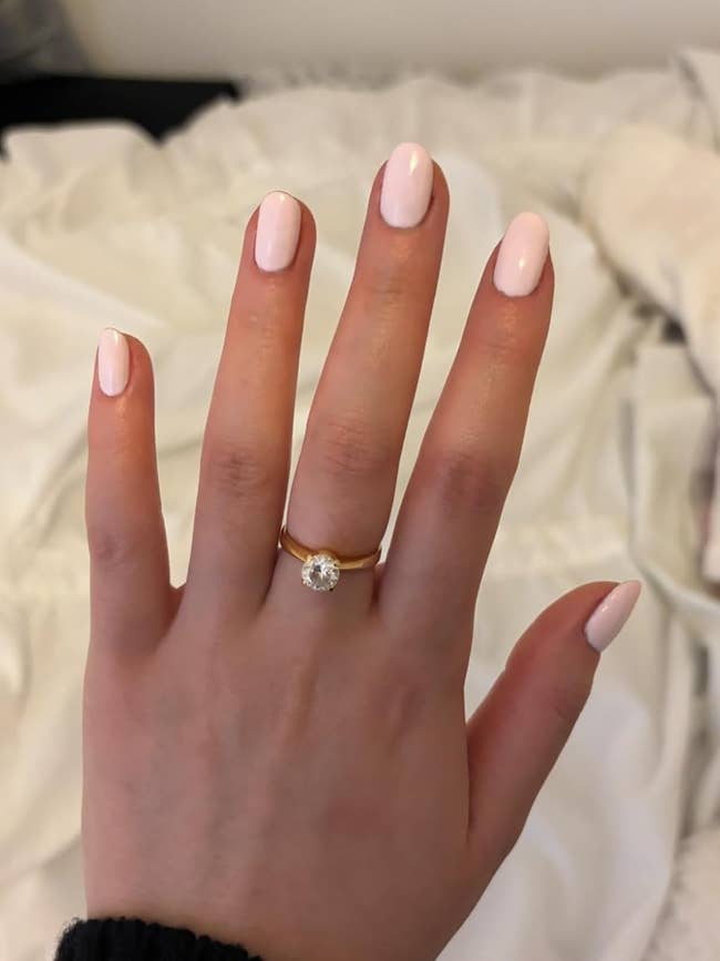 A hand with light pink manicured nails displays a gold ring featuring a large round diamond, set against a softly blurred background