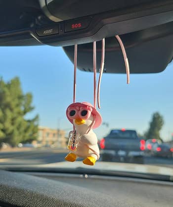 A small duck figurine in a white robe and pink sunglasses hanging from a car's rearview mirror