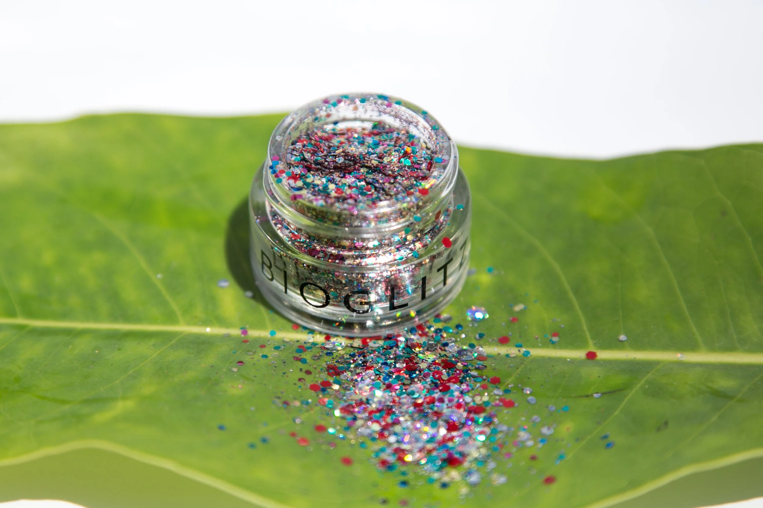 A tiny  jar of multicolored glitter sits connected  a leaf, with glitter spilling out, suggesting a vibrant, eco-friendly cosmetic product